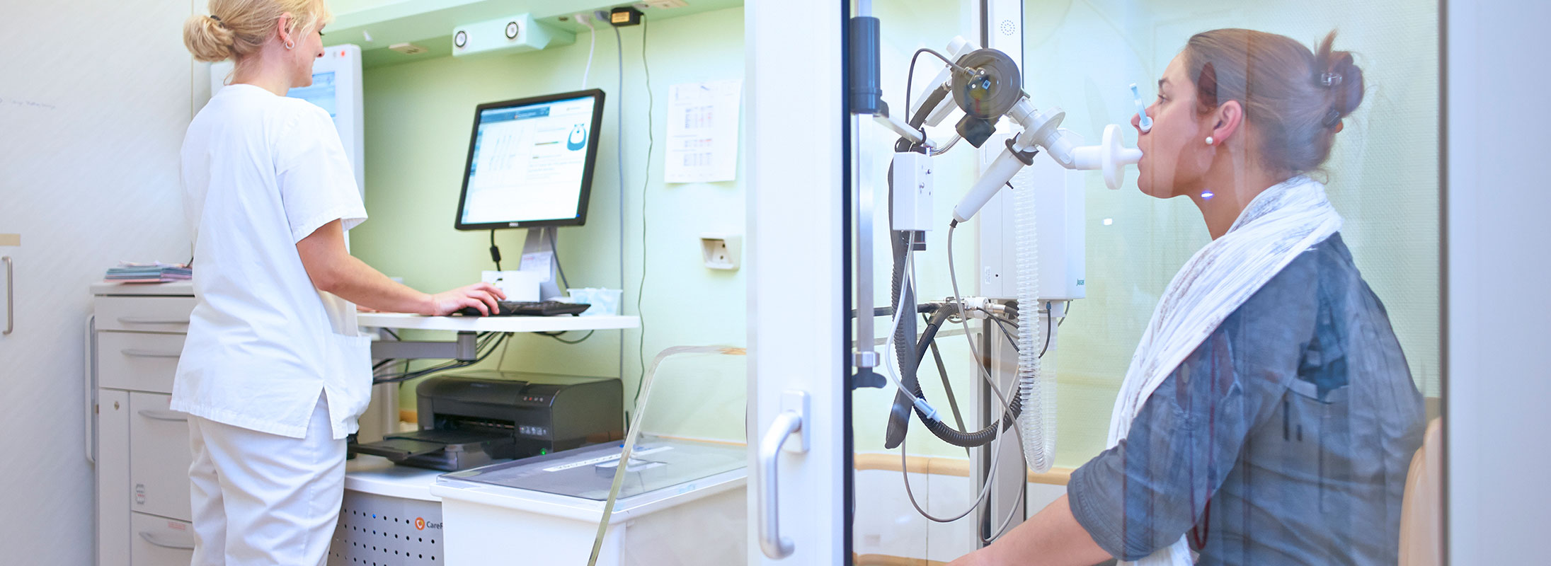 Medizinische Fachangestellte betreut eine Patientin bei einer Lungenfunktionsmessung in einer Kabine.
